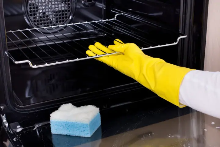Main tenant porte-gants jaunes pour nettoyer le four. Une éponge bleue et blanche est posée sur le fond du four.