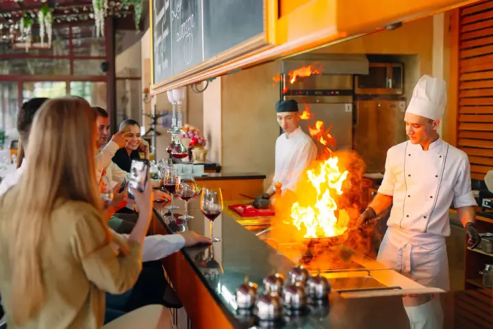 Cuisine ouverte d'un restaurant, chefs en action avec des flammes impressionnantes. Des clients prennent des photos, respectant la norme.