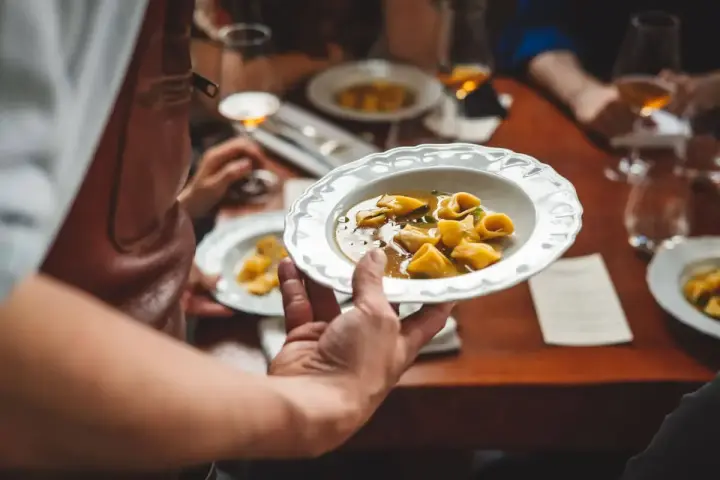 Un serveur présente une assiette de tortellinis dans un bouillon. Le service en restauration est à son apogée.