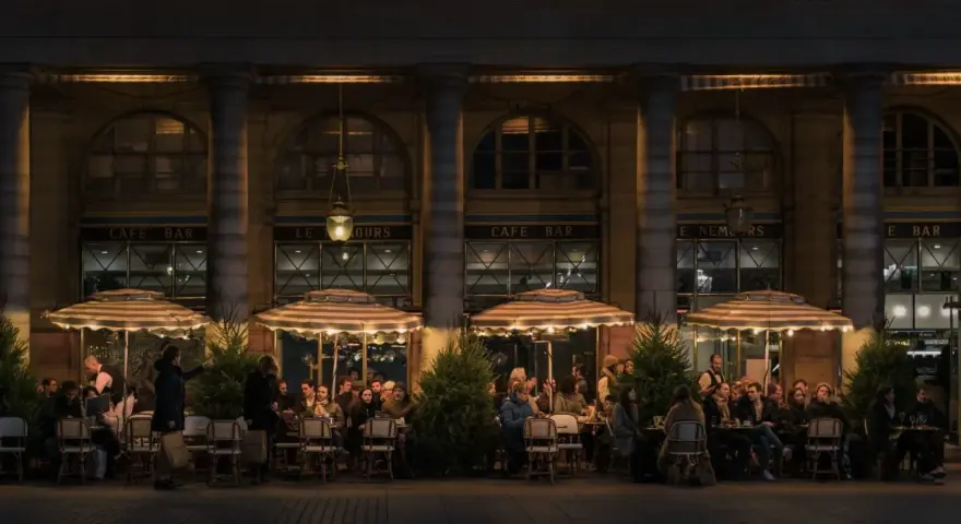 Terrasse de café animée à **heure de nuit**, éclairée par des lampes et des parasols rayés. Des clients discutent et boivent, profitant de l'ambiance parisienne.