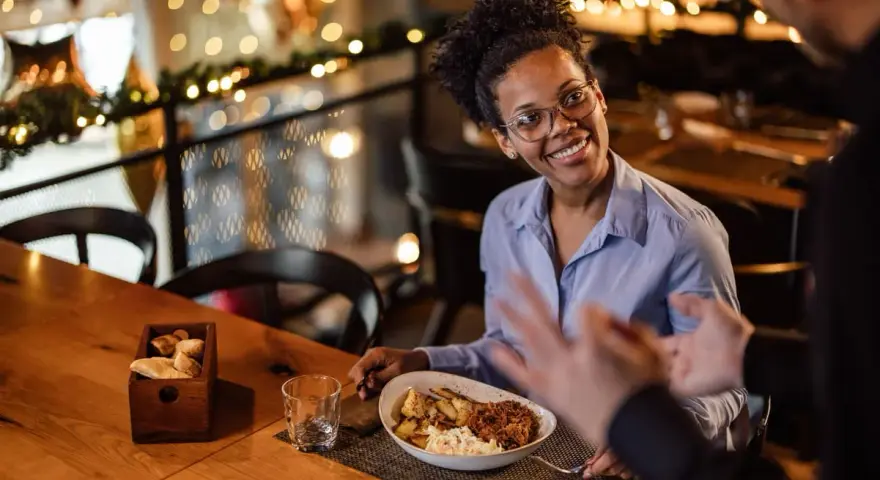Une femme souriante discute avec un homme, son assiette de nourriture devant elle. Elle semble partager son avis sur la cuisine, une véritable critique culinaire.
