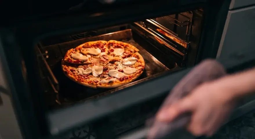 Une main avec une manique retire une pizza du four. La question est : quelle température pour réchauffer une pizza au four ?