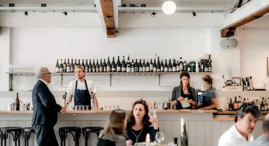 Un bar de restaurant animé avec un personnel attentif, des bouteilles de vin alignées et des clients profitant de l'ambiance. La gestion du restaurant assure un service fluide.