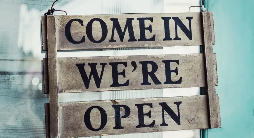 Texte annonce ouverture restaurant : panneau en bois "COME IN WE'RE OPEN" accroché à une porte vitrée.