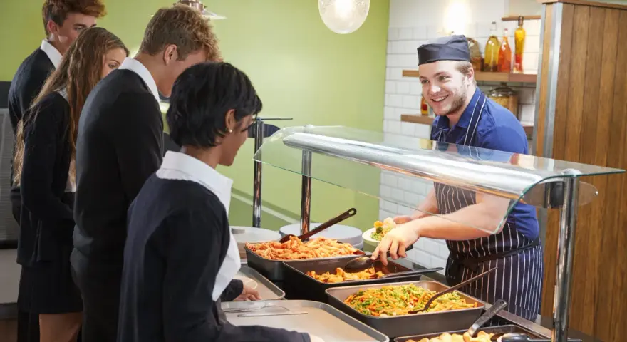 Des élèves font la queue pour le déjeuner, attendant leur plat. Le cuisinier, souriant, sert des légumes. Un contrat d'usage est peut-être en jeu pour ce repas scolaire.