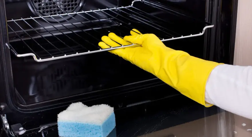 Main tenant porte-gants jaunes pour nettoyer le four. Une éponge bleue et blanche est posée sur le fond du four.