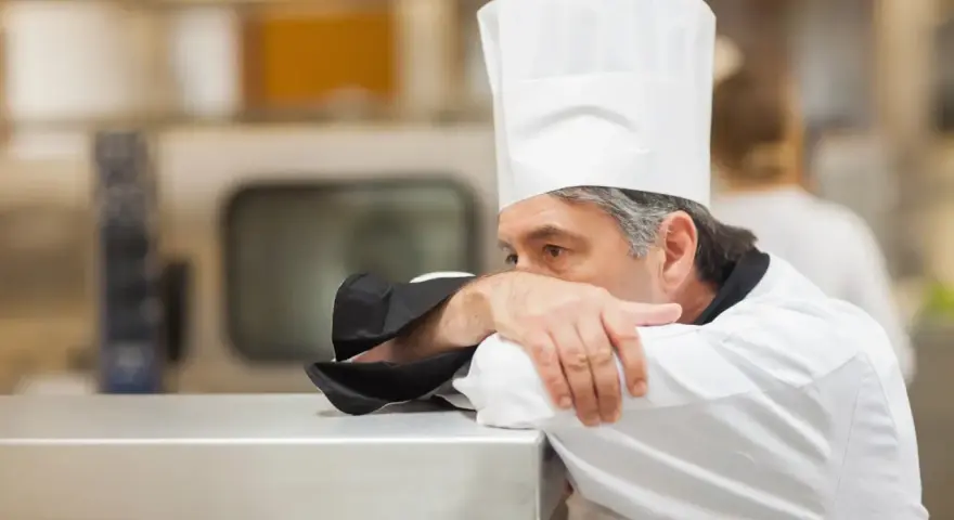 Un chef fatigué, le visage caché dans son bras, semble épuisé par un long jour de carence restauration rapide.