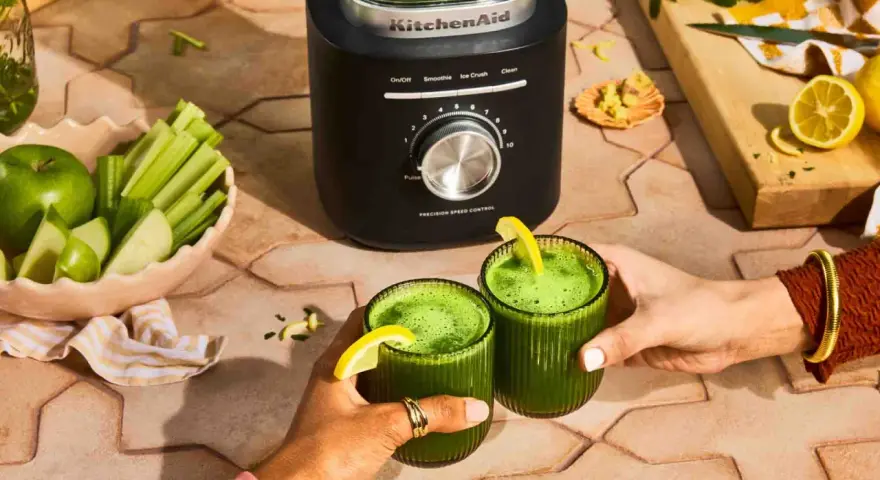 Deux mains portent des verres de jus vert, préparé avec un blender KitchenAid. Pommes, céleri et citrons sont prêts à être mixés.