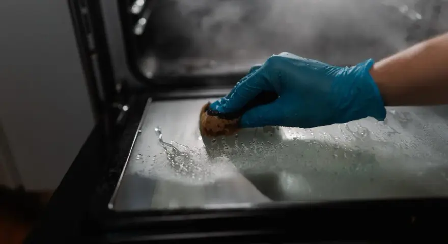 Main gantée bleue pour nettoyer le four avec du bicarbonate. La vapeur s'échappe, signe que le bicarbonate a chauffé.