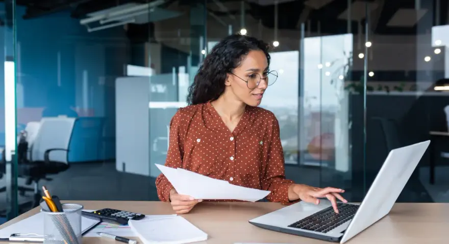 Femme travaillant sur un ordinateur portable, tenant des documents. Elle semble réfléchir à une décision unilatérale de l'employeur.