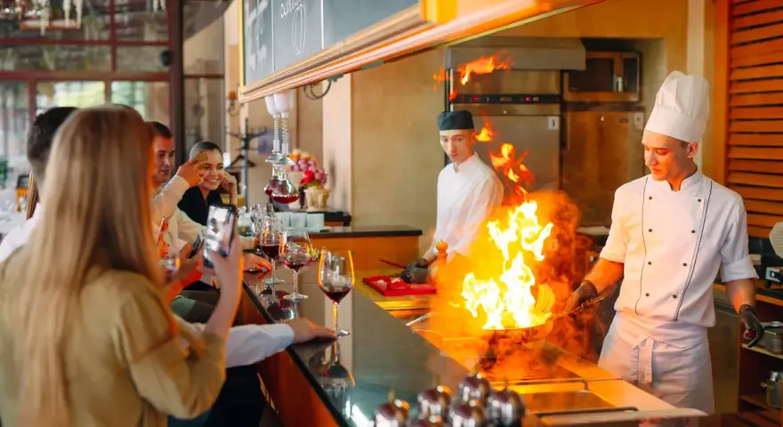 Cuisine ouverte d'un restaurant, chefs en action avec des flammes impressionnantes. Des clients prennent des photos, respectant la norme.