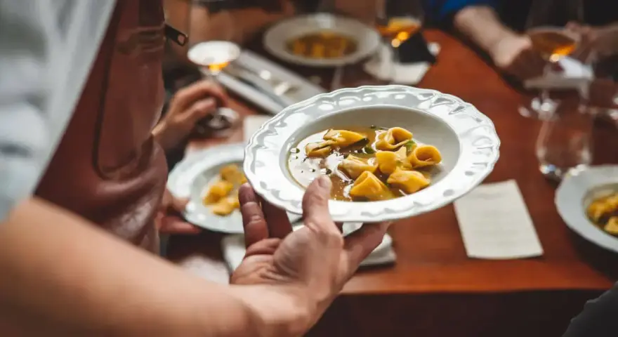 Un serveur présente une assiette de tortellinis dans un bouillon. Le service en restauration est à son apogée.