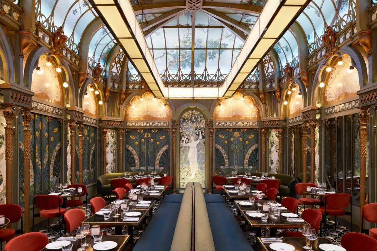 Magnifique restaurant Art Nouveau avec des banquettes bleues, des chaises rouges velours et des décorations florales dorées sous un plafond de verre.