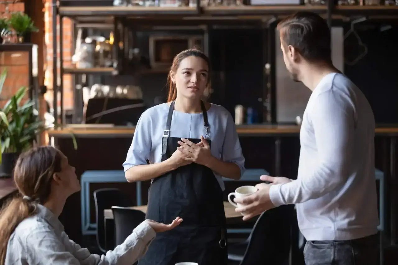 Serveuse attentive face &agrave; une r&eacute;ponse client m&eacute;content. Elle &eacute;coute avec empathie, les mains jointes sur son tablier.