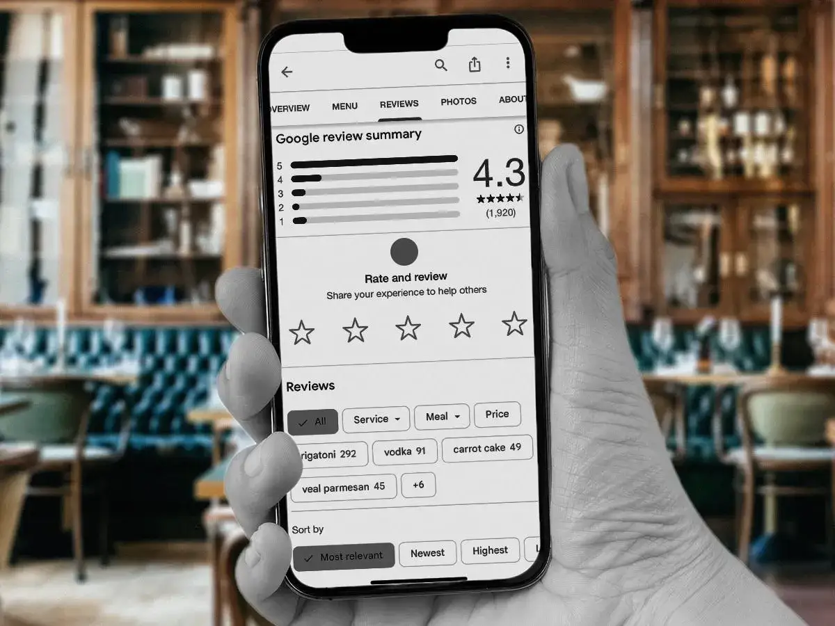 Main tenant dans un restaurant, un smartphone affiche l'interface pour laisser un avis Google. La note moyenne est de 4.3 étoiles.