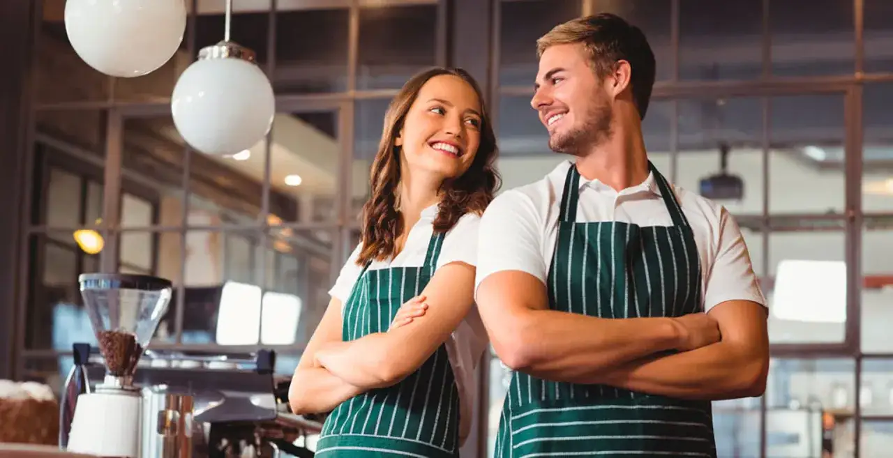 Deux jeunes employés souriants, un homme et une femme, portant des tabliers verts rayés dans un café.