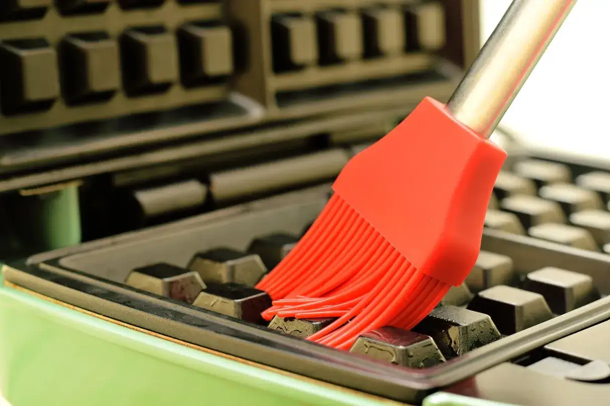 Un pinceau rouge nettoie un gaufrier encrassé. Les poils en silicone atteignent les recoins pour un nettoyage parfait.