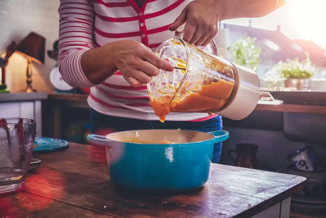 Une personne verse une purée orange d'un blender dans une cocotte bleue. C'est à quoi sert un blender : préparer des purées onctueuses.