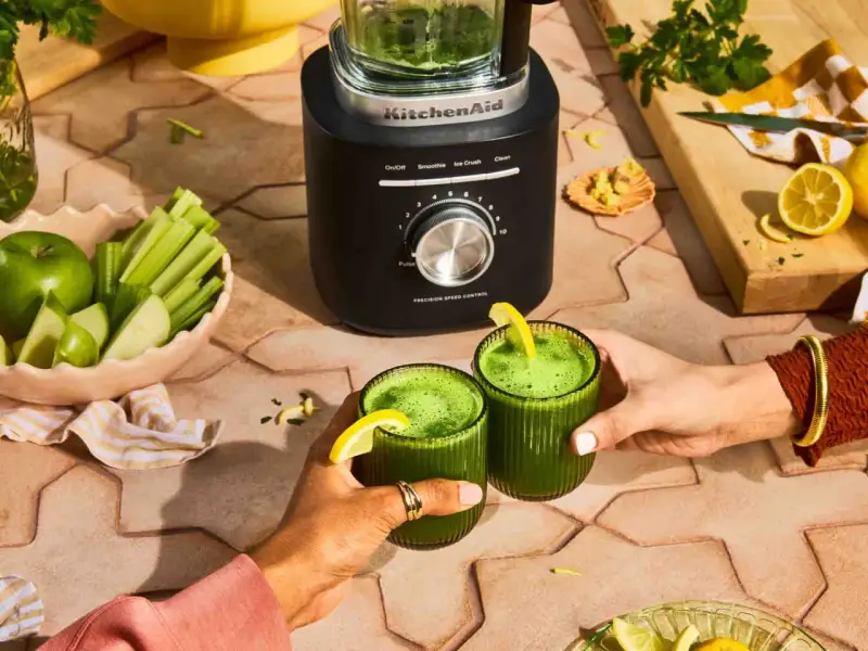 Deux mains portent des verres de jus vert, préparé avec un blender KitchenAid. Pommes, céleri et citrons sont prêts à être mixés.